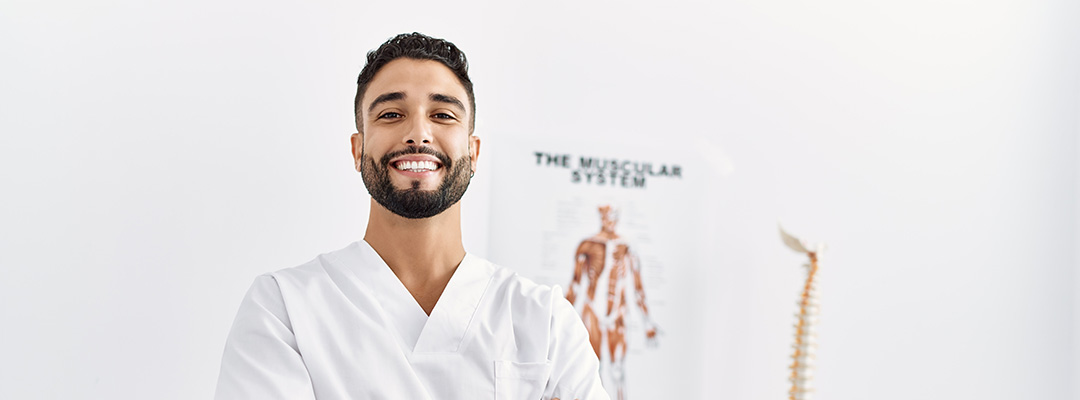 Young physiotherapist standing with arms crossed gesture at clinic with models behind him.